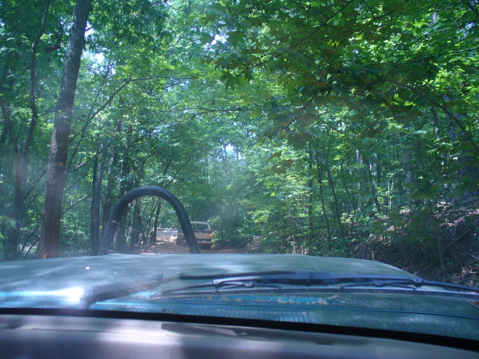 View from inside a vehicle looking down an off-road trail with trees on both sides. Two other vehicles are visible in the distance