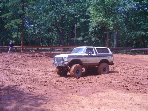 1996 Ford Bronco - A lifted Ford Bronco navigates a muddy terrain with dense woods visible behind a fence.