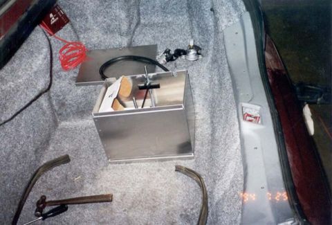 1992 Ford Crown Victoria - A polished aluminum battery box with a hold-down rod and top plate mounted to the gray-carpeted trunk floor of a car, alongside a red batte…