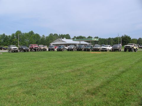 1995 Ford C-350 (Centurion Conversions) - Several modified off-road vehicles are parked in a line on a sunny day near a gas station.