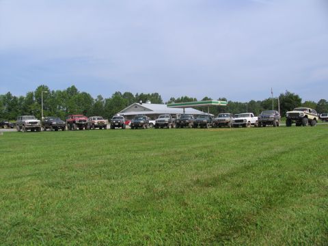 1995 Ford C-350 (Centurion Conversions) - A group of off-road vehicles are parked in a grassy lot in front of a building and gas station.
