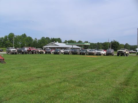 1995 Ford C-350 (Centurion Conversions) - A collection of modified trucks and SUVs are gathered in a grassy field near a gas station.