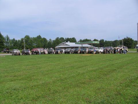 1995 Ford C-350 (Centurion Conversions) - A collection of trucks and SUVs are gathered in a field, with a gas station and trees in the background.
