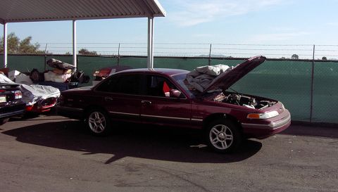 1992 Ford Crown Victoria - Maroon Ford Crown Victoria sedan with the hood up and a cover draped over the fender, parked under a metal carport canopy in a fenced lot b…