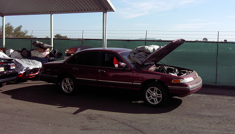 Maroon Ford Crown Victoria with hood up under a carport in a fenced storage lot