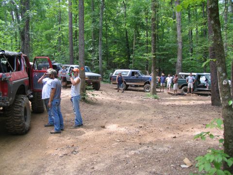 1995 Ford C-350 (Centurion Conversions) - A group of off-road vehicles are parked in a wooded area, with several people standing around them.
