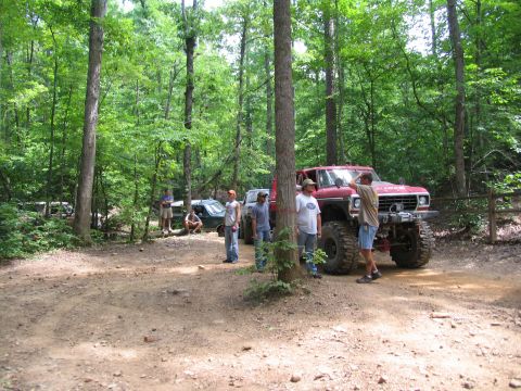 1995 Ford C-350 (Centurion Conversions) - Several people gather around a heavily modified red pickup truck in a forest clearing.