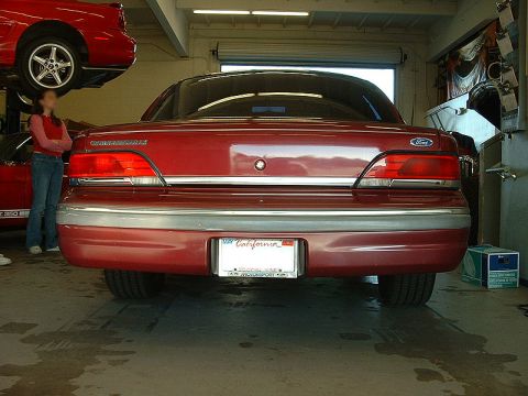 1992 Ford Crown Victoria - Straight-on rear view of a dark red aero-body Ford Crown Victoria LX inside a shop, showing wraparound taillights with chrome trim, a Ford…