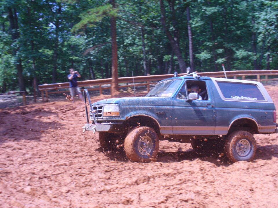 Blue 1996 Ford Bronco driving through mud with a person taking a photo in the background