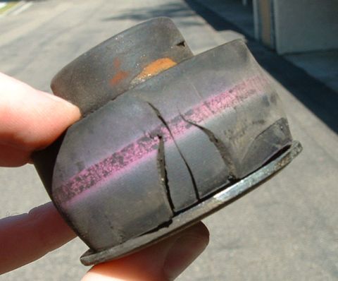 1992 Ford Crown Victoria - A hand holds a cylindrical rubber-and-metal suspension bushing that has cracked and split apart, with visible pink/purple wear banding and…