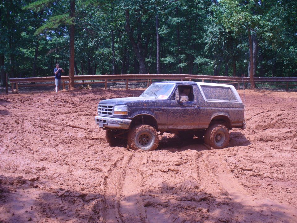Dark blue Ford Bronco stuck in mud, 3/4 front view. Muddy tires and body. Trees and a person in background