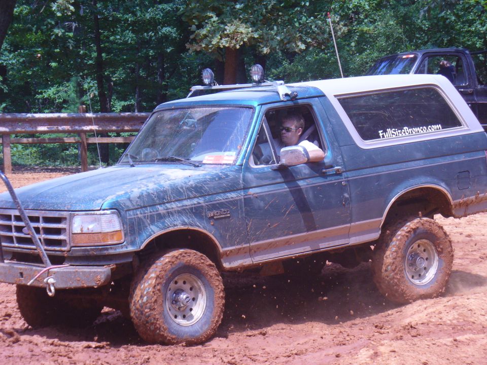 Green 1996 Ford Bronco with white top driving on a muddy off-road trail. Another truck is visible behind it