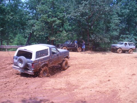 1996 Ford Bronco - Multiple SUVs, including a muddy Ford Bronco, navigate a dirt and mud track surrounded by trees.