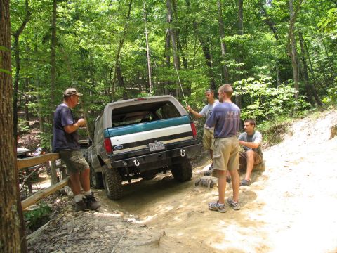 1995 Ford C-350 (Centurion Conversions) - A group of men gather around a Ford Bronco on a wooded off-road trail.