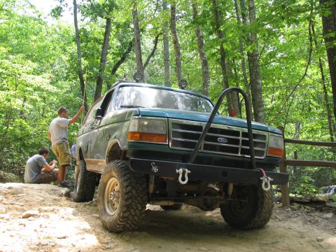 1995 Ford C-350 (Centurion Conversions) - A modified Ford Bronco sits on a rocky trail surrounded by lush green trees.