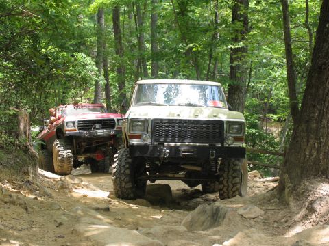 1995 Ford C-350 (Centurion Conversions) - Two lifted Ford trucks navigate a rocky, wooded trail during an off-road event.