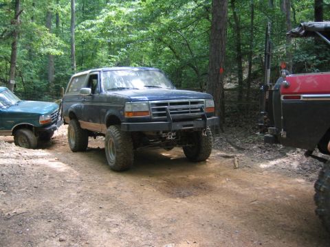 1979 Ford Bronco - Two off-road SUVs are parked on a dirt path surrounded by trees.