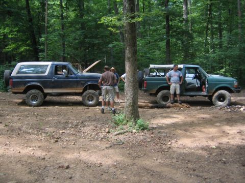 1979 Ford Bronco - Two modified SUVs, a dark blue Bronco and a green pickup, are parked side-by-side on a dirt path in a wooded area.