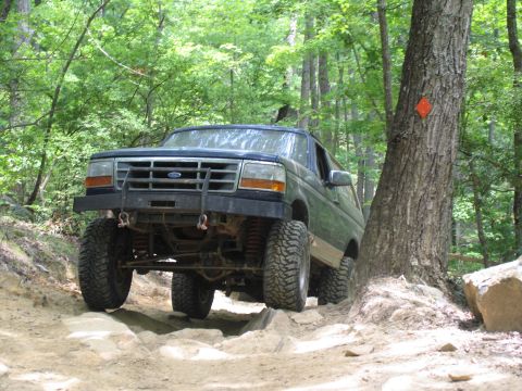 1995 Ford C-350 (Centurion Conversions) - A lifted Ford Bronco tackles a challenging, rocky path surrounded by dense trees.
