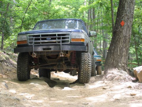 1995 Ford C-350 (Centurion Conversions) - Two Ford trucks drive on a wooded trail, one in front with large tires and a bull bar.