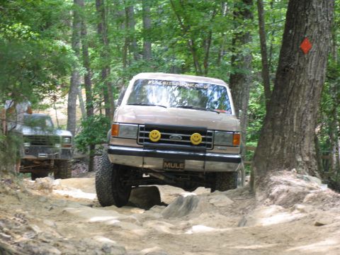 1995 Ford C-350 (Centurion Conversions) - A tan Ford Bronco drives over rocks on a wooded trail, with another vehicle visible behind it.