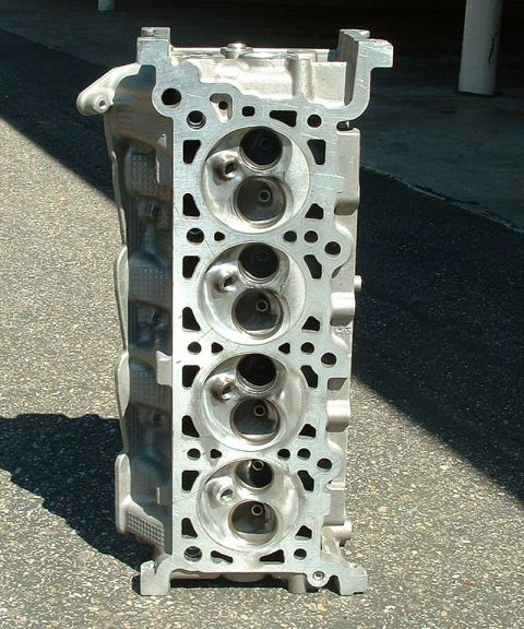 1992 Ford Crown Victoria - Bare aluminum cylinder head standing upright on sunlit asphalt, combustion-chamber side facing the camera, showing four chambers with insta…
