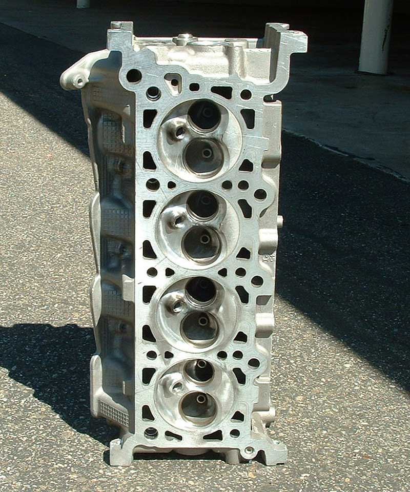 Bare aluminum four-chamber cylinder head standing on pavement, valves installed