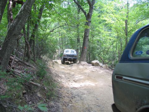 1995 Ford C-350 (Centurion Conversions) - A dark Ford F-Series truck navigates a rocky dirt path through a wooded area, with another vehicle visible in the foreground.