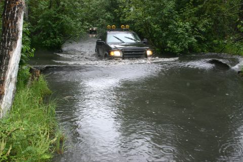 2003 Jeep TJ - A dark SUV navigates a water crossing surrounded by dense foliage.