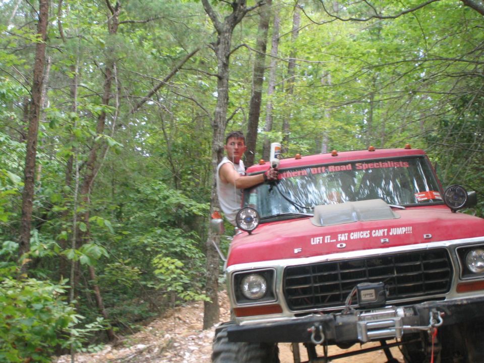 Man leaning on the hood of a red lifted Ford truck with winch on a wooded trail