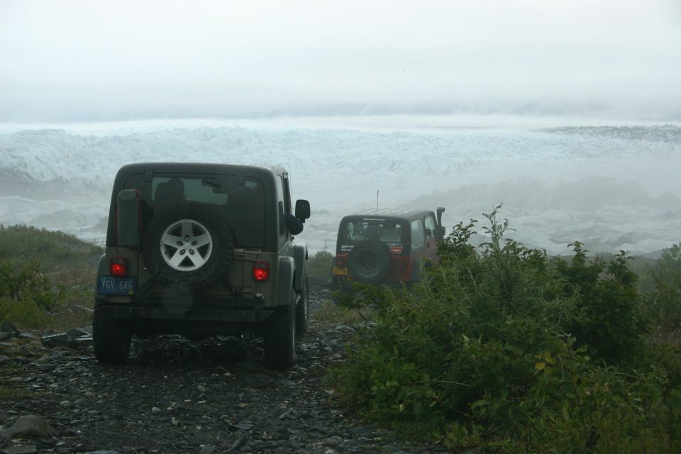 Two Jeep TJs on a gravel road, one dark green, one red, with a glacier in the background