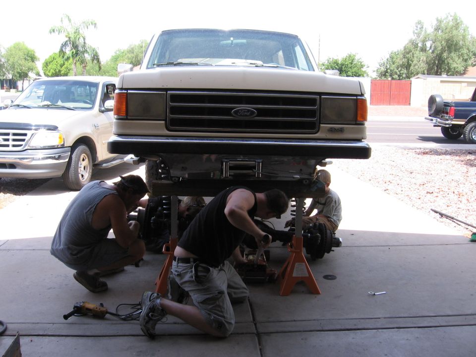 People working under a lifted 1987 Ford Bronco. Another Ford pickup is parked nearby