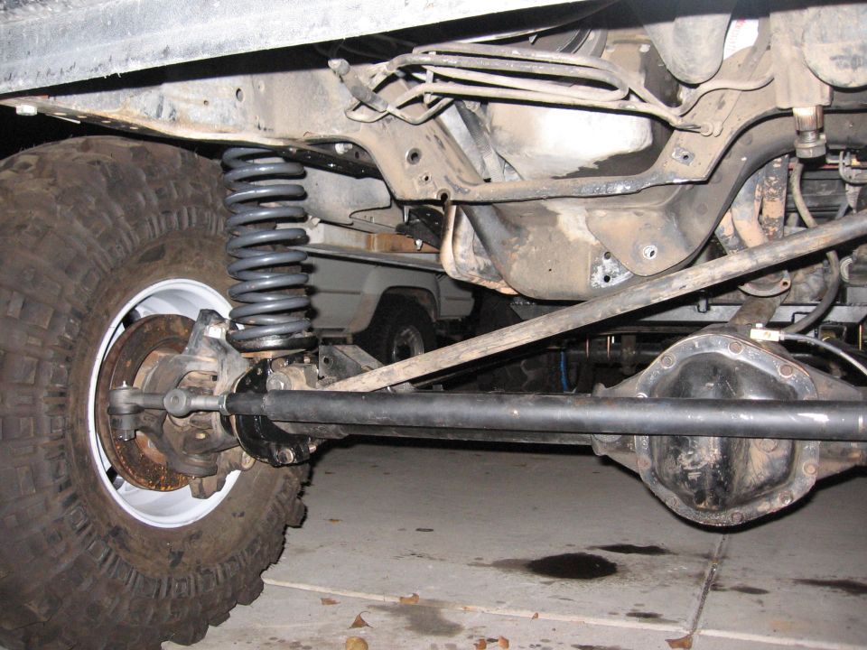 Undercarriage view of a vehicle's front axle and suspension with a large tire. Another vehicle is partially visible in the background