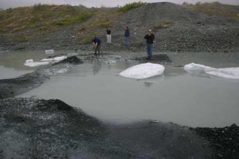 2003 Jeep TJ - People explore a glacial meltwater area with ice floes and dark, rocky terrain under an overcast sky.