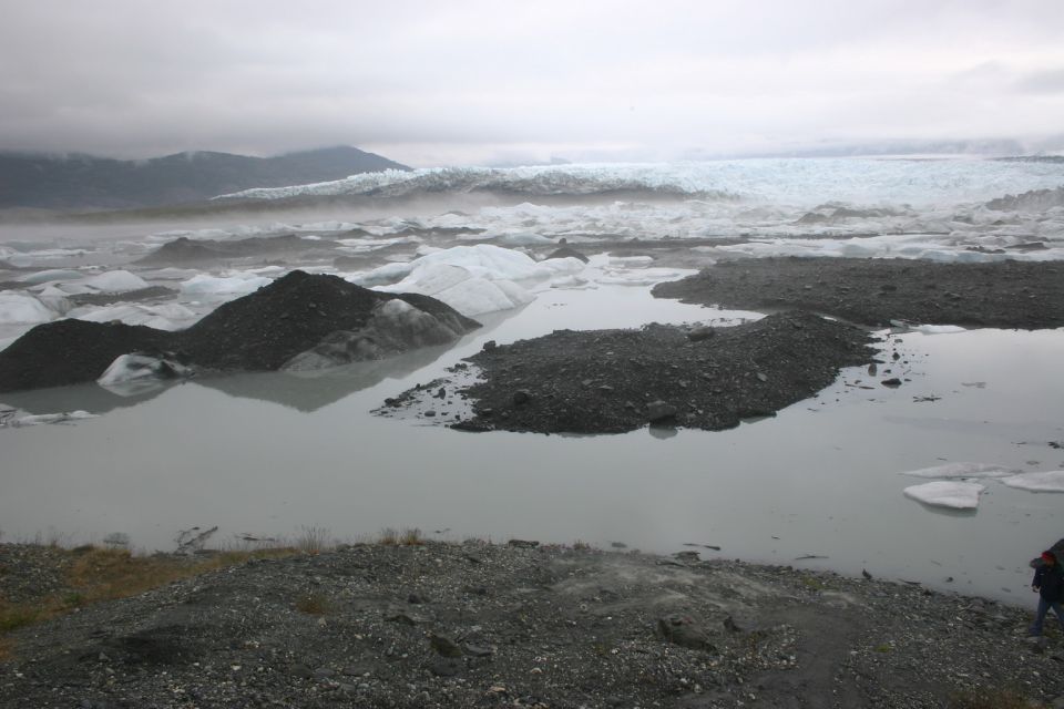 Glacier in background with icebergs floating in a lake, foreground has gravel and a person on the right