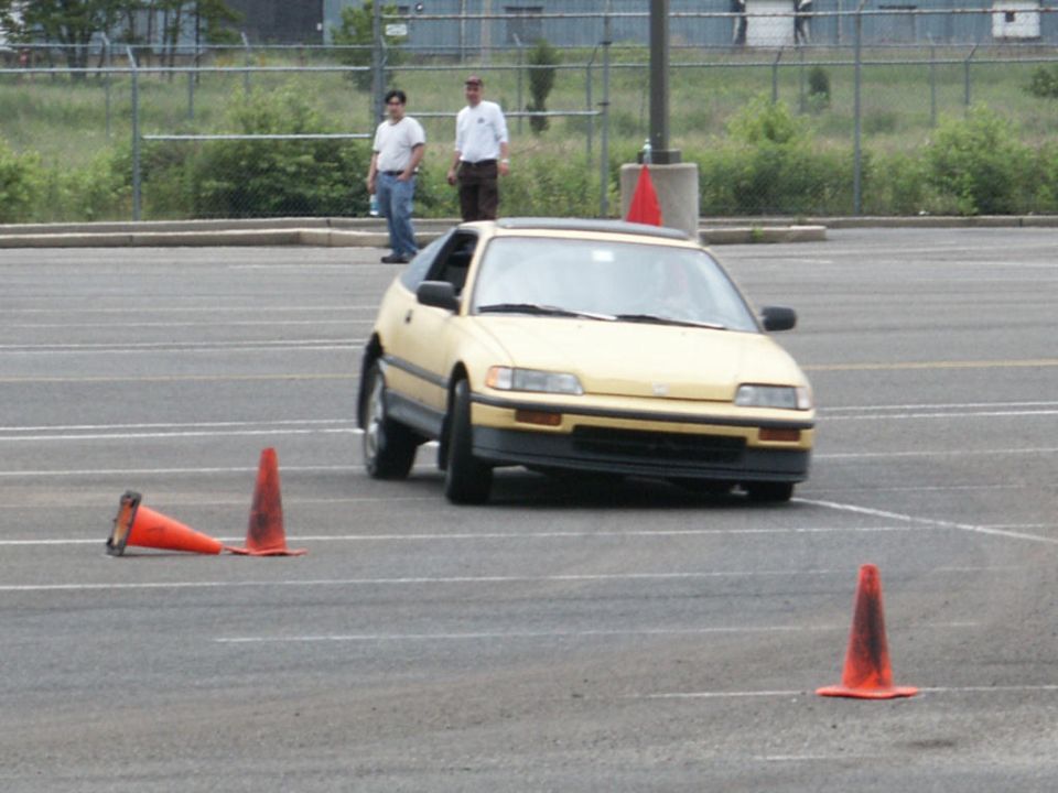 Yellow hatchback car navigating a course marked by orange cones. Two men stand in the background