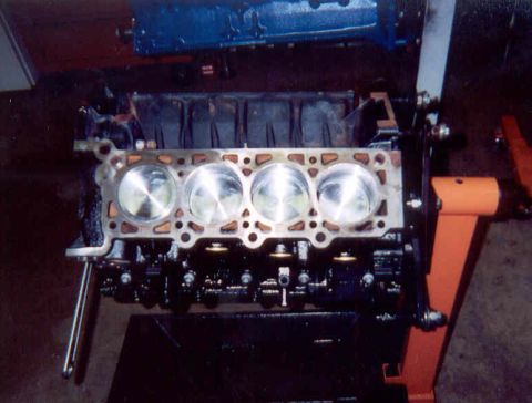 1992 Ford Crown Victoria - Top-down view of one bank of a V8 short block on an orange engine stand, with a head gasket laid over the deck and four polished flat-top p…