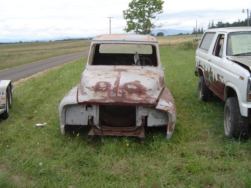 Rusty stripped 1955 Ford F-100 cab without grille or fenders in a grassy field, Bronco parked beside it