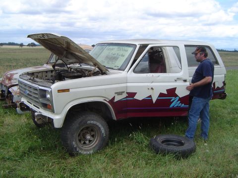 1955 Ford F-100 - A white Ford Bronco sits in a grassy field with its hood open, a man stands by the driver's door, and a spare tire rests on the grass nearb…
