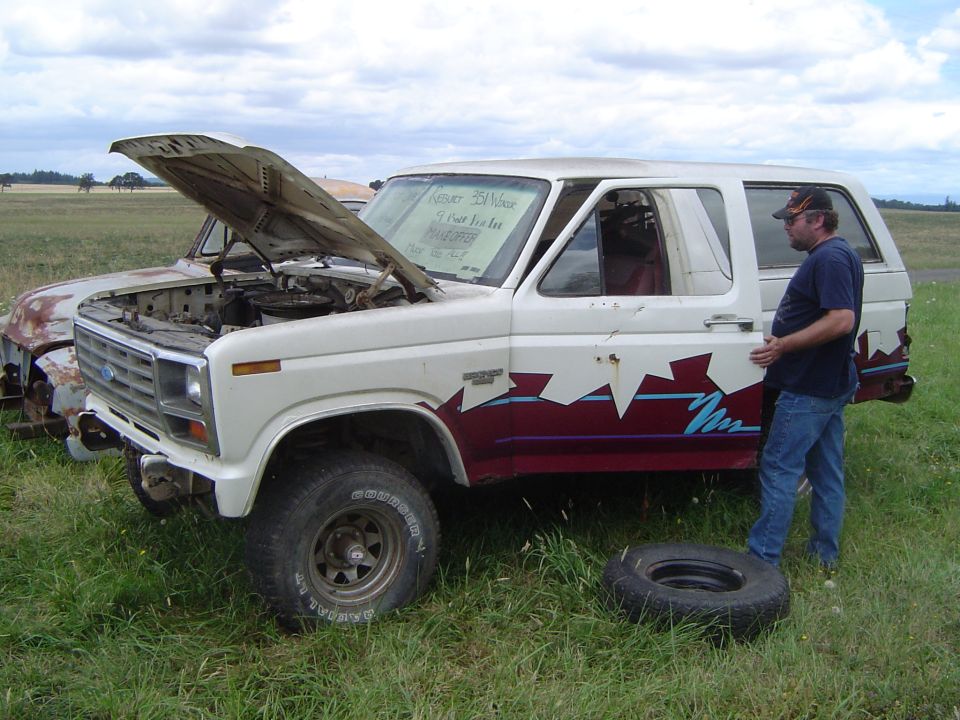 White Ford Bronco with open hood, man standing by door, spare tire on ground
