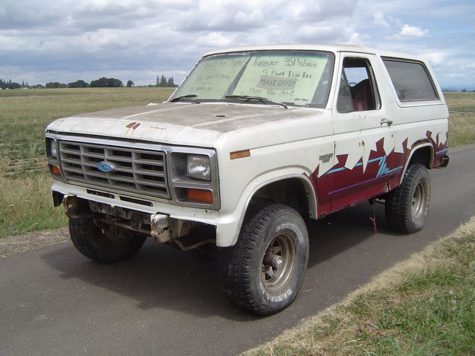 White Ford Bronco with red and blue graphics on the side. Windshield has signs for sale. Front bumper is damaged
