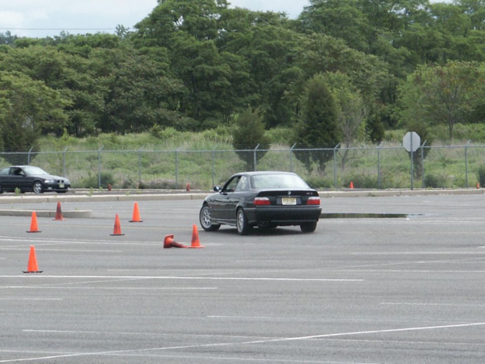 Black BMW 3 Series coupe driving on a paved lot with orange cones; another car is visible in the background