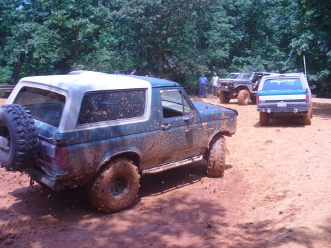 1996 Ford Bronco - Several Ford Broncos are seen off-roading on a muddy trail surrounded by trees.
