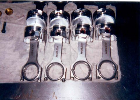 1992 Ford Crown Victoria - Four forged pistons pressed onto polished Manley H-beam connecting rods, laid out in a row on a white cloth over a workbench. The rod caps…