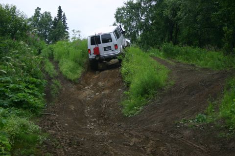 2003 Jeep TJ - A white van navigates a challenging off-road trail, with lush green foliage surrounding the muddy path.