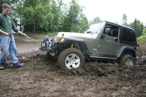 2003 Jeep TJ - A person attempts to pull a stuck olive green Jeep TJ out of deep mud with a winch cable.