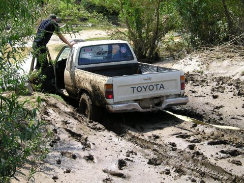 1993 Ford Bronco - A white Toyota pickup truck is stuck deep in mud, with a person attempting to assist from the driver's side door.