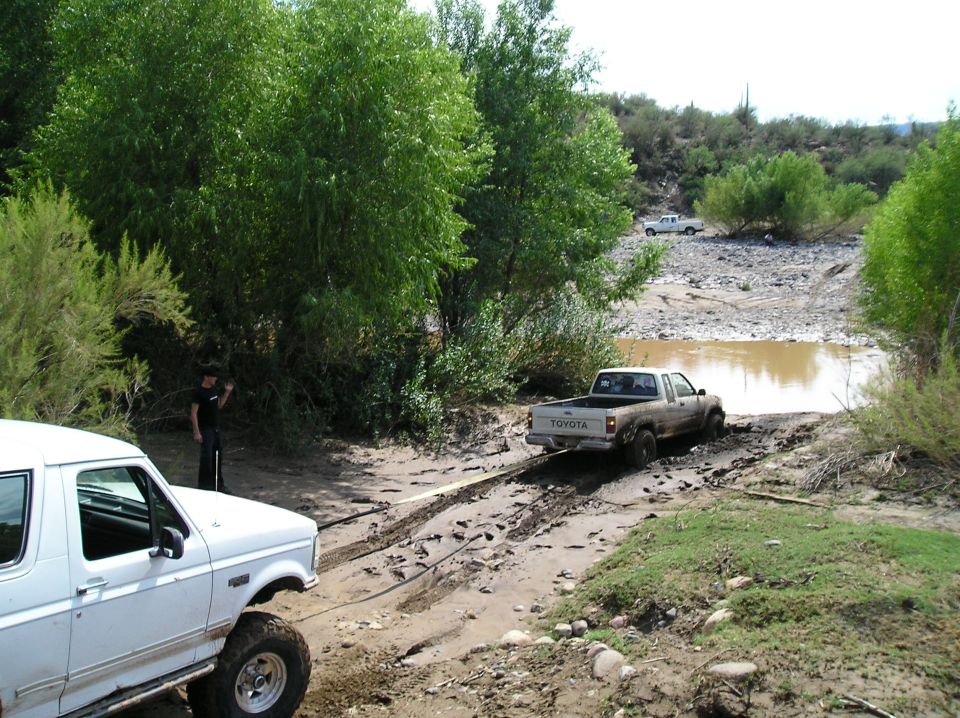 White Ford Bronco on lifted tires towing a stuck Toyota pickup at a muddy desert creek crossing
