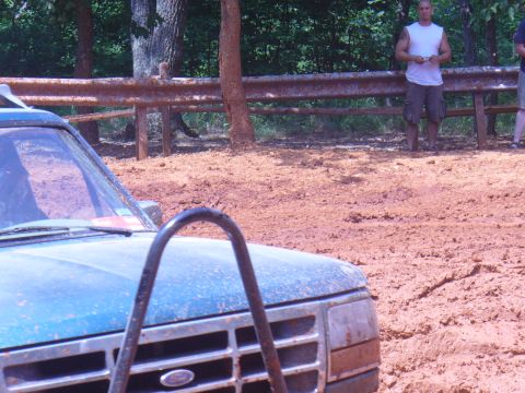 1996 Ford Bronco - A blue Ford truck is stuck in thick mud, with two onlookers standing behind a rusty guardrail.