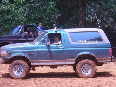 1996 Ford Bronco - A green 1996 Ford Bronco is seen off-road with muddy tires, parked next to another truck.